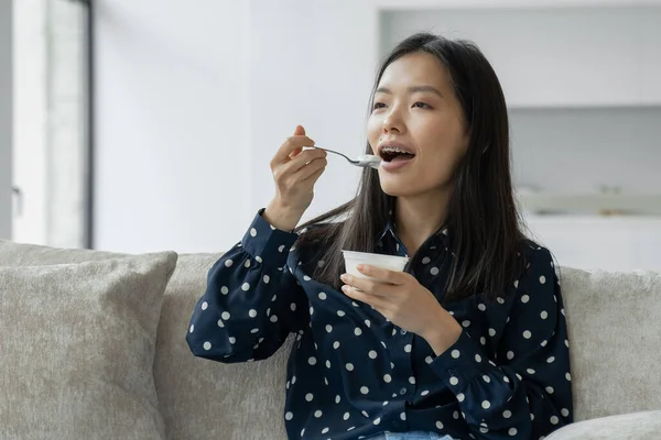 Joven mujer asiática come yogur fresco para el desayuno sentado en el sofá — Foto de Stock