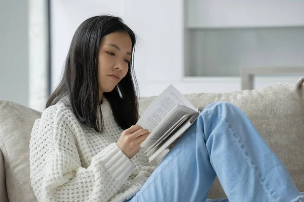 Mujer asiática feliz en un suéter blanco sentado en el sofá y leyendo un libro en casa — Foto de Stock