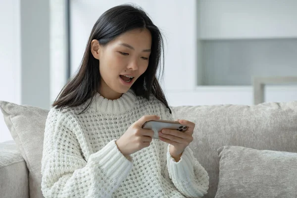 Una joven asiática emocionada está sentada en el sofá de la sala de estar, jugando un juego móvil en un teléfono inteligente — Foto de Stock