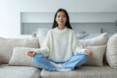Oturma odasında yoga yapan Asyalı kız kanepede oturuyor. Lotus pozisyonunda oturan dişi bir yogi. Yoga ve meditasyon kavramı