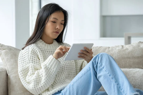 Mulher asiática usa um tablet enquanto se senta em um sofá em casa em sua sala de estar. Uma mulher feliz usa um tablet para assistir a vídeos e navegar na internet em casa. — Fotografia de Stock