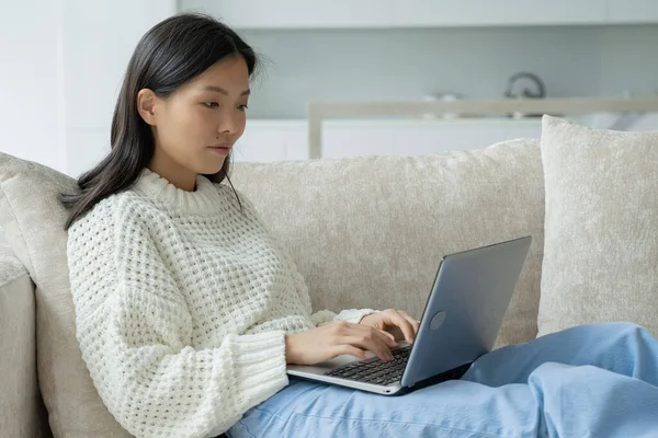 En una sala de estar moderna, una mujer asiática con un ordenador portátil está sentada en el sofá. Una mujer se dedica a un trabajo remoto, navegar por la web — Foto de Stock