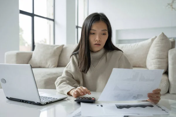 Mulher asiática com um laptop e uma calculadora na sala de estar. Uma senhora séria com a ajuda de um computador portátil calcula as finanças em casa e olha para suas contas no papel — Fotografia de Stock