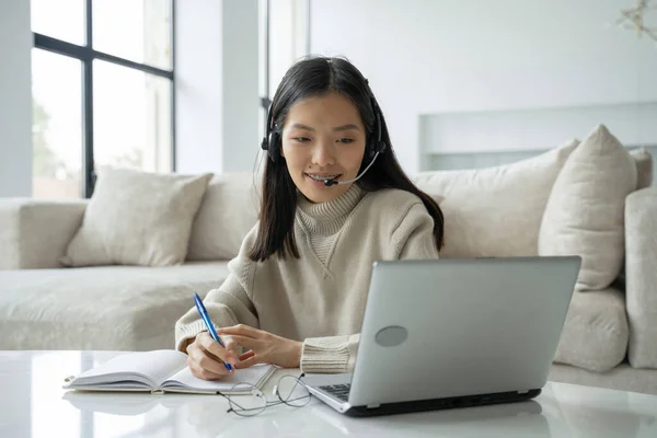 Uma mulher asiática usando um fone de ouvido olha para a tela do laptop, se comunica com o interlocutor. Mulher de negócios coreana com fones de ouvido chamada de vídeo para atendimento ao cliente — Fotografia de Stock