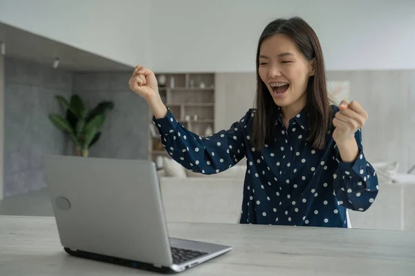 Emocionada joven asiática alegre se sienta en una mesa con un ordenador portátil, levanta las manos y celebra el éxito de la victoria — Foto de Stock
