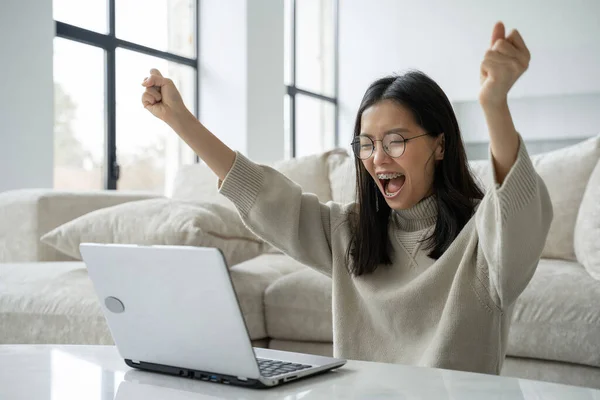 Una exitosa empresaria asiática con gafas disfruta del éxito con un portátil en su escritorio. Una mujer alegre que ha recibido un premio mayor celebrando su victoria. — Foto de Stock