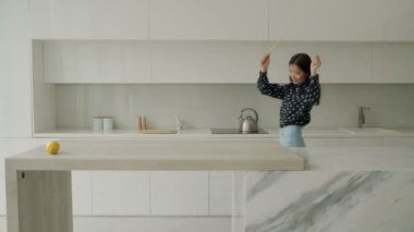 Gülümseyen, tişörtlü, kot pantolonlu güzel bir Asyalı kadın modern bir mutfakta dans ediyor, sağlıklı yemekler pişiriyor mutlu bir kadın sevdiği müziğe geçiyor.