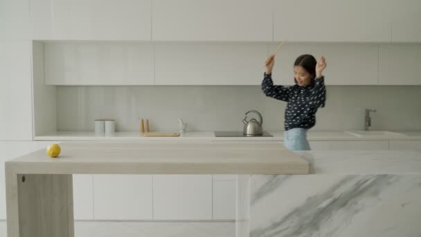 Sonriendo hermosa mujer asiática en una camisa y jeans baila en una cocina moderna, cocina comida saludable una mujer feliz se mueve a su música favorita — Vídeo de stock