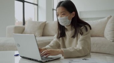Maskeli düşünceli bir Asyalı kadın oturma odasında dizüstü bilgisayar kullanıyor. Öğrenci çevrimiçi çalışıyor, uzaklık öğreniyor