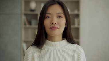 Asyalı kadın mutlu hissediyor, gülümsüyor ve oturma odasında duran kameraya bakıyor.