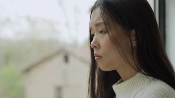 Een jonge teleurgestelde Aziatische vrouw zit in het huis en kijkt uit het raam. — Stockvideo