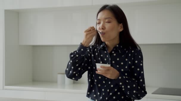 若いアジアの女性は、キッチンで朝食のために新鮮なヨーグルトを食べる — ストック動画