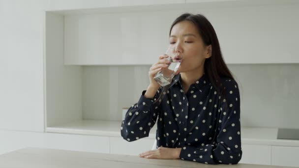 若いアジアの女性は台所に座っているガラスから水を飲む。家で水を飲みながら笑顔を見せる若い女性。ライフスタイルコンセプト. — ストック動画