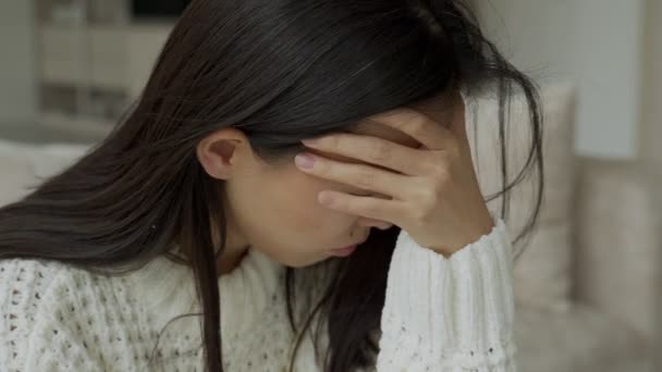 Mão sobre os templos de uma jovem mulher asiática triste sentada no sofá. Ela sente-se mal por causa da dor de cabeça. — Vídeo de Stock