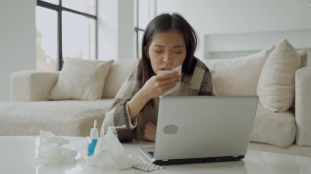 An Asian woman blows her nose in a napkin, sits at a home office desk, studies on a laptop, a sick woman has the flu, covered herself with a blanket, caught a cold — Stock Video