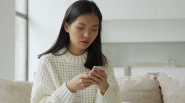 Oturma odasındaki kanepede akıllı telefon kullanan güzel bir Asyalı kadının portresi.