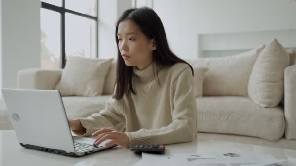 リビングルームでラップトップと電卓を持つアジアの女性。ラップトップコンピュータの助けを借りて深刻な女性は、家の財政を計算します。. — ストック動画