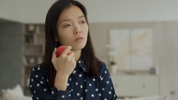 Nachdenkliche Asiatin hält einen Apfel im Wohnzimmer und schaut aus dem Fenster — Stockvideo