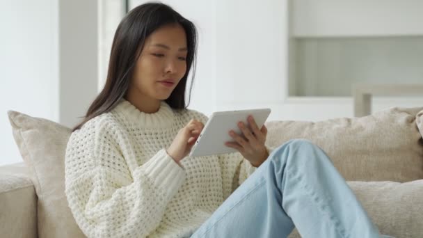 リビングルームでソファに座っている間、アジアの女性はタブレットを使用しています。幸せな女性はビデオを見て、自宅でインターネットをサーフィンするためにタブレットを使用しています. — ストック動画