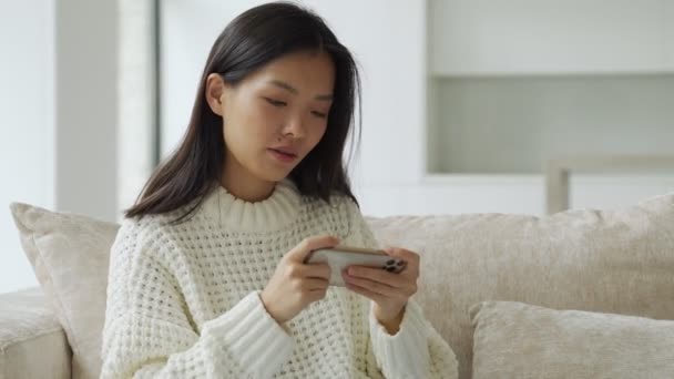 興奮した若いアジアの女性がリビングルームのソファに座っていて、スマートフォンでモバイルゲームをしています。 — ストック動画