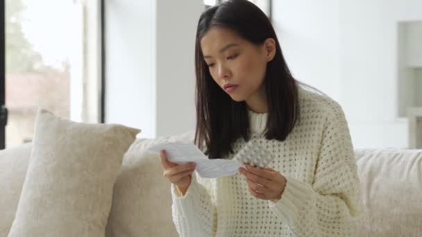 Femme asiatique avec un rhume et la grippe lit des instructions avec des pilules dans ses mains, assis sur le canapé — Video