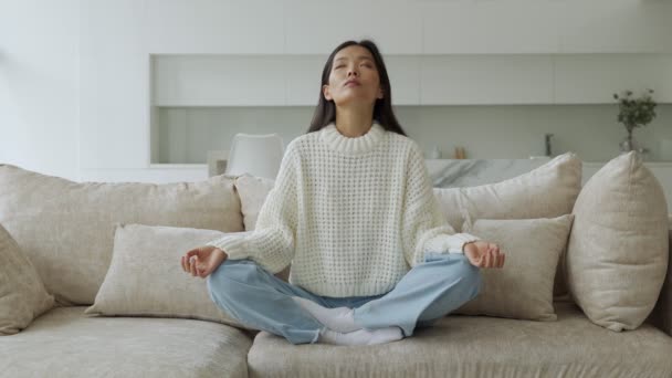 Ázsiai lány jógázik a nappaliban a kanapén ülve. Egy női jógi lótuszpozícióban. A jóga és a meditáció fogalma — Stock videók