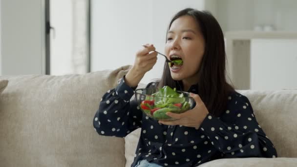 Νεαρή ευτυχισμένη Ασιάτισσα που τρώει υγιεινή σαλάτα καθισμένη στον καναπέ στο σαλόνι. — Αρχείο Βίντεο