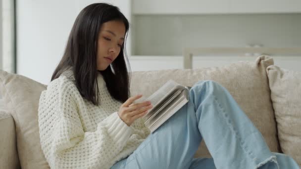 Heureuse femme asiatique dans un pull blanc assis sur le canapé et lisant un livre à la maison — Video