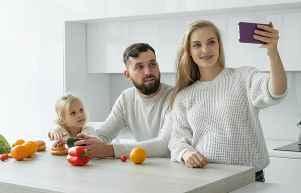 Gelukkig familie leven, een zwangere vrouw schiet een video blog op een smartphone met haar man en dochtertje in de keuken — Stockfoto