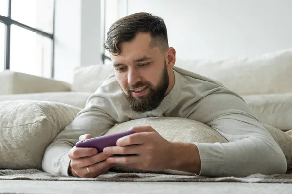Portret van een opgewonden jonge man met een baard die op de vloer ligt en spelletjes speelt op een mobiele telefoon — Stockfoto