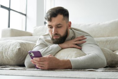 Sakallı genç adam evin zemininde akıllı telefon kullanıyor.