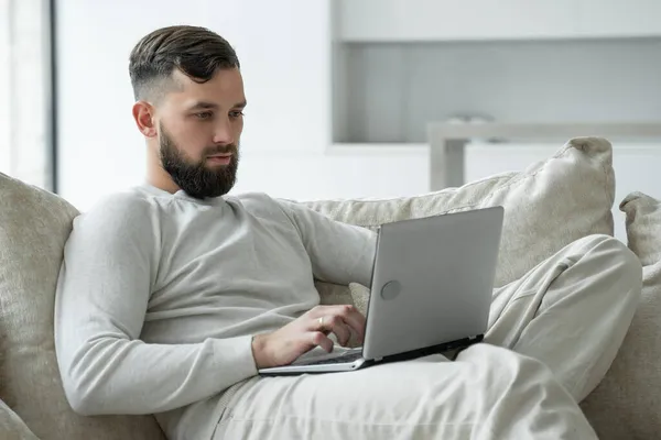 Hombre barbudo joven utiliza un ordenador portátil para trabajar en casa en la sala de estar — Foto de Stock