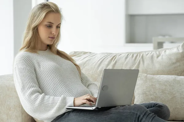Mujer embarazada está sentada en un sofá beige con un ordenador portátil. El concepto de una mujer embarazada que trabaja en casa — Foto de Stock
