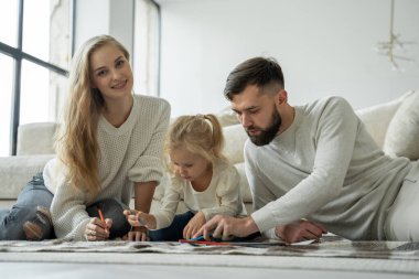 Genç bir aile, küçük sevimli bir kızı olan evli bir çift, modern bir evde yerde yatıyorlar, kağıda çiziyorlar, renkli kalemler kullanıyorlar.