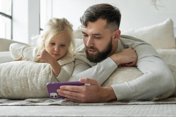 Baard vrolijke vader en zijn dochtertje kijken video 's op een smartphone en lachen vrolijk liggend op de vloer in de woonkamer — Stockfoto