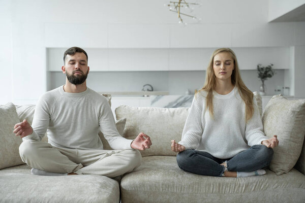 Calm young family is doing yoga together, sitting in the lotus position on the sofa. Parents relax on the couch, meditating
