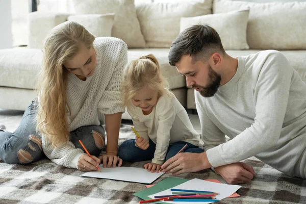 Jonge familie, een getrouwd stel met een kleine charmante dochter, liggend op de vloer in een modern huis, tekent op papier, gebruikt kleurpotloden — Stockfoto