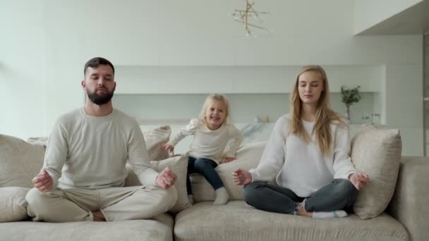Los padres jóvenes mamá y papá están sentados en casa en el sofá, meditando, haciendo yoga, alivia las emociones negativas. Energetic pequeña hija salta en el sofá y se interpone en el camino de sus padres — Vídeo de stock