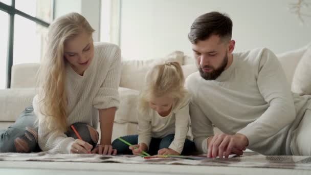 Família jovem, um casal com uma pequena filha encantadora, deitada no chão em uma casa moderna, desenha no papel, usa lápis de cor — Vídeo de Stock