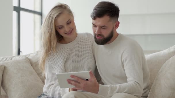 Giovane coppia felice, guardando lo schermo del tablet, rilassarsi su un comodo divano. Una coppia innamorata fa acquisti online in un negozio online. — Video Stock