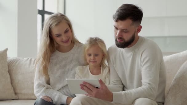 Mutlu bir aile evde, oturma odasındaki kanepede otururken eğleniyor, ebeveynler ve küçük bir kız dijital tablet kullanıyorlar, ekrana bakıyorlar, kanepede birlikte oturuyorlar, gülüyorlar. — Stok video