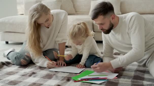 Junge Familie, Ehepaar mit kleiner charmanter Tochter, liegt in einem modernen Haus auf dem Boden, zeichnet auf Papier, verwendet Buntstifte — Stockvideo