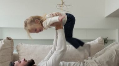 Sakallı baba kanepede uzanıyor ve küçük kızıyla oynuyor. Onu kollarında tutuyor. Babası kızını kollarında tutuyor, çocuk babasının kollarında uçuyor..