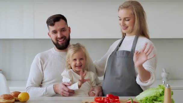 Portrait of a young couple with a little daughter standing indoors in the kitchen at home, smiling and looking at the camera and waving their hands — Stock Video