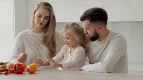Glückliche Familie kocht zusammen in der Küche. Eine schwangere Mutter zeigt ihrer kleinen Tochter Gemüse und Obst. Erholung zu Hause und Kochen am Wochenende — Stockvideo