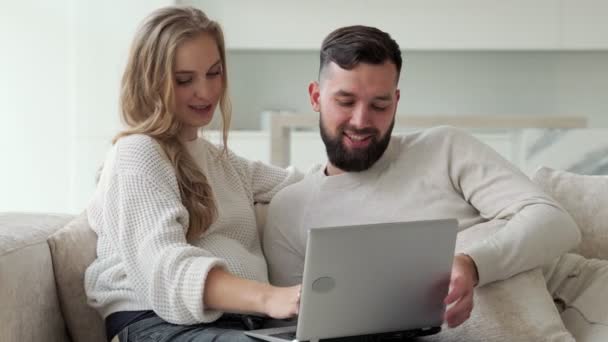 Feliz jovem casal está usando um laptop juntos, sentado em casa em um sofá aconchegante, um homem e uma mulher estão olhando para uma tela de computador — Vídeo de Stock