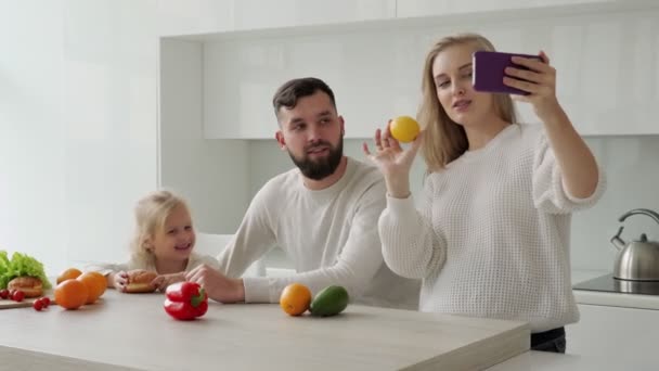 Lycklig familj lever, en gravid kvinna fotograferar en video blogg på en smartphone med sin man och lilla dotter i köket — Stockvideo