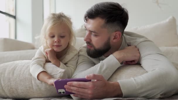 Skäggig glad pappa och hans lilla dotter tittar på videor på en smartphone och skrattar glatt liggande på golvet i vardagsrummet — Stockvideo