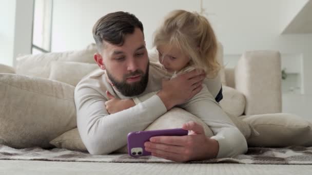 Бородатый веселый папа и его маленькая дочь смотрят видео на смартфоне и весело смеются, лежа на полу в гостиной — стоковое видео