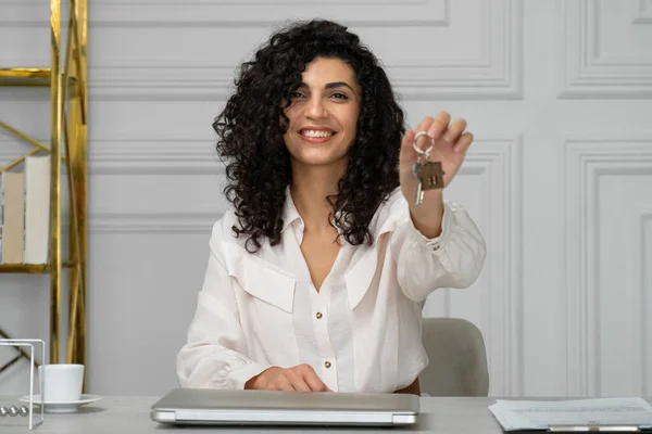 Индийская женщина с вьющимися волосами, агент по недвижимости или брокер, показывает ключи от нового дома или квартиры, сидя за столом в офисе — стоковое фото